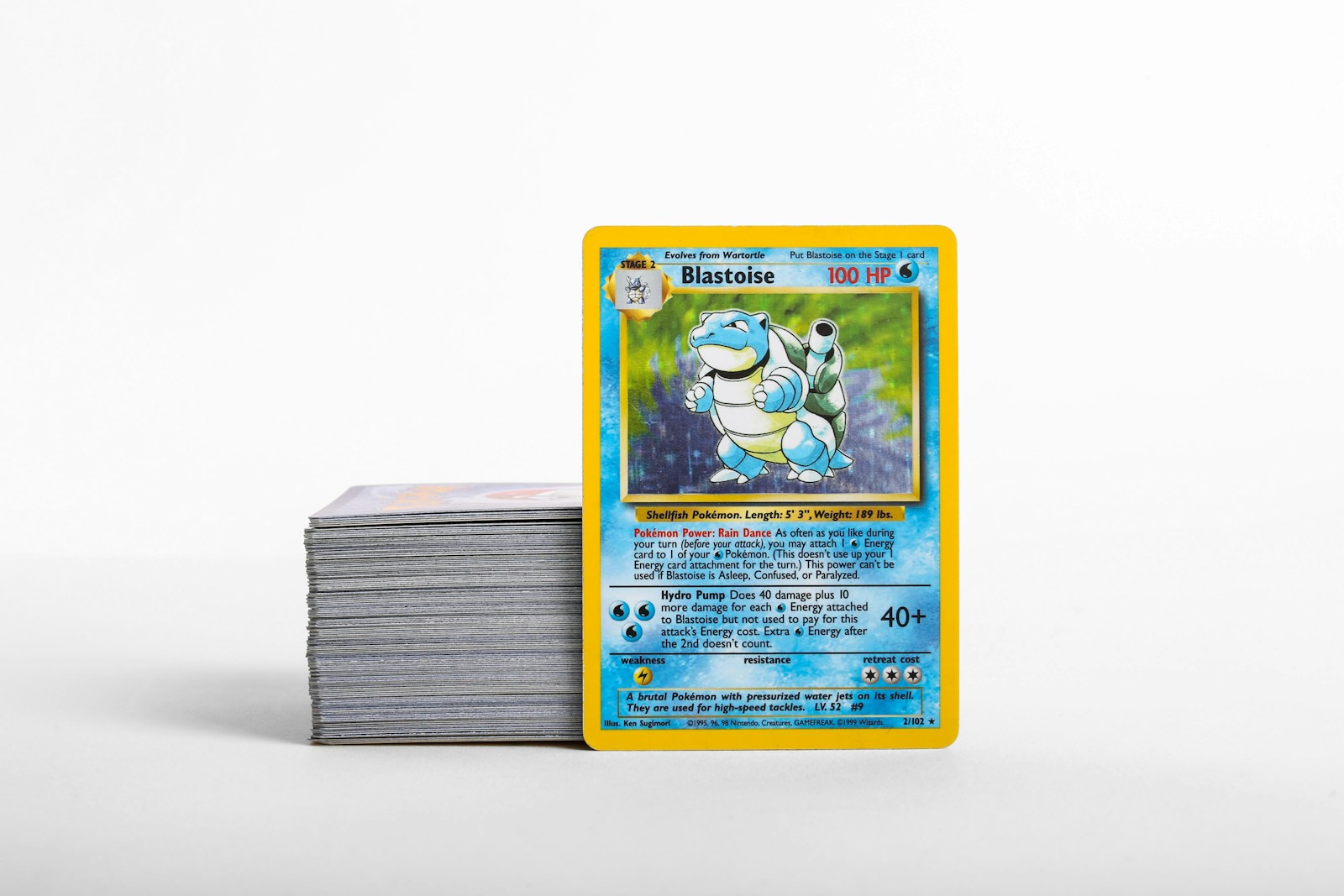 Une carte Pokémon Blastoise à côté d’une pile de cartes à collectionner sur une surface claire.